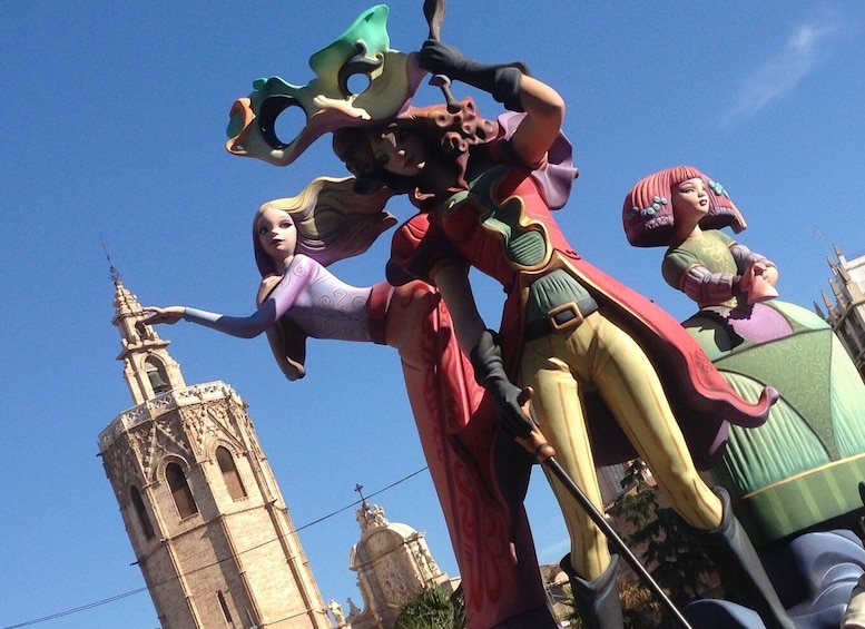 Colorful sculptures representing characters from Valencia's Fallas festival, with a historic tower in the background under a clear blue sky.