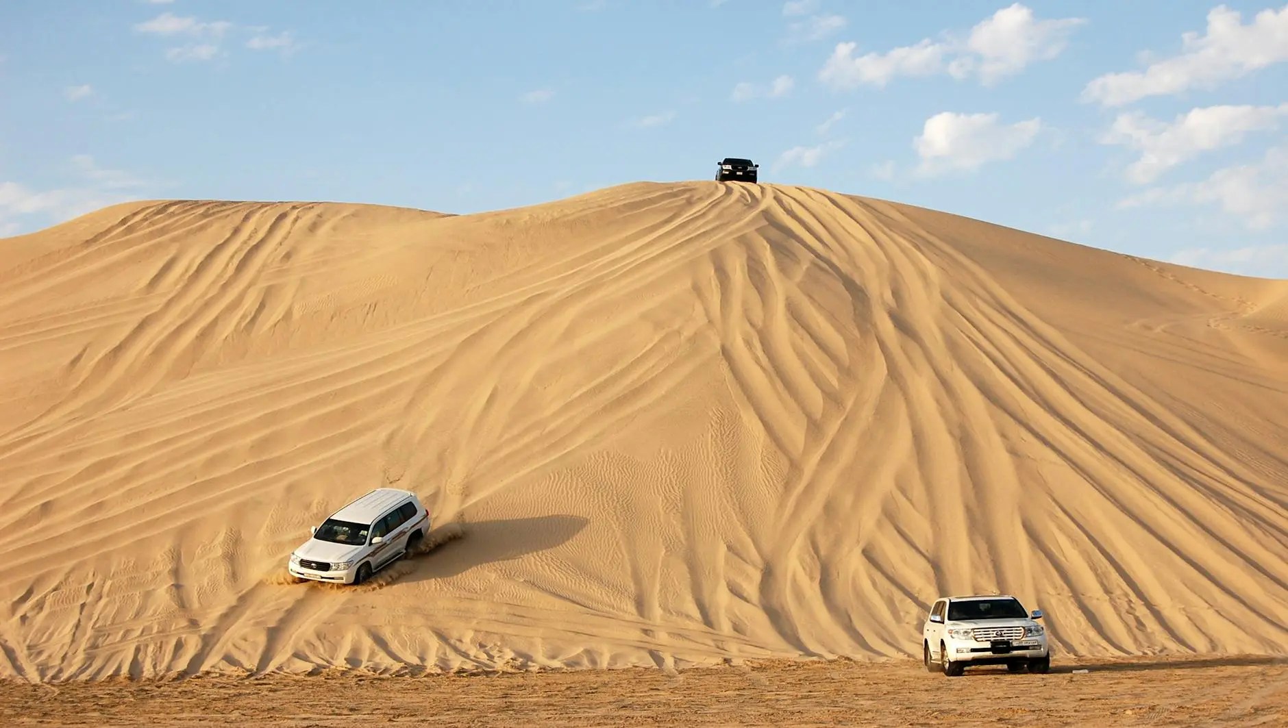 Two SUVs driving on sand dunes under a blue sky with clouds.
