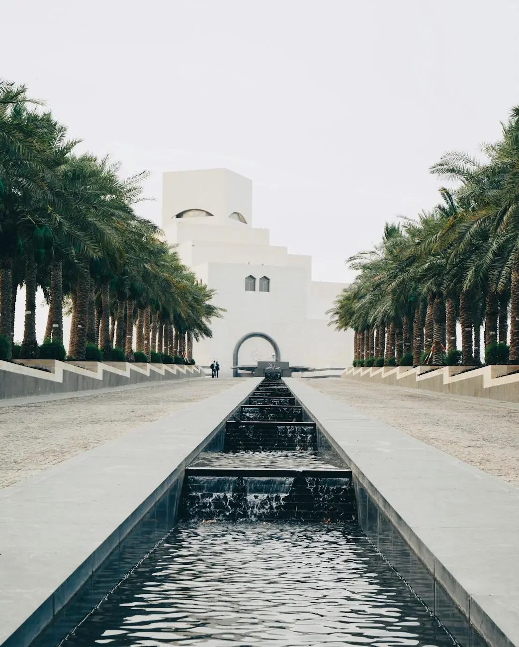 A view of the Museum of Islamic Art in Doha, Qatar, framed by palm trees and a pathway with a water feature leading towards the building.