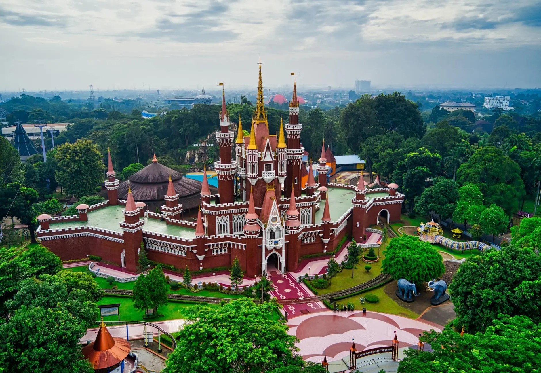 Aerial view of a large, red-bricked castle with tall spires and surrounded by green trees and gardens, indicating a theme park setting.