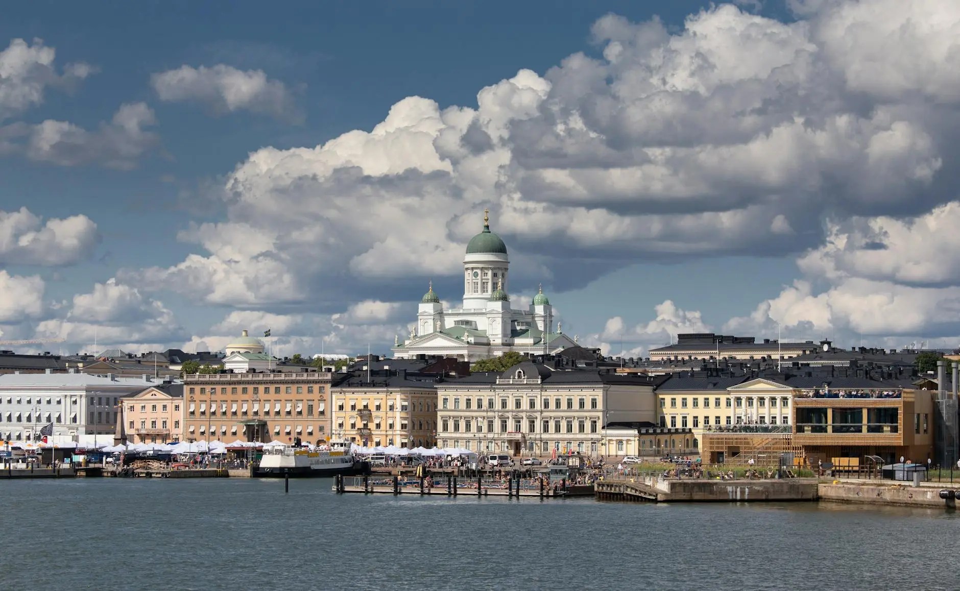 A panoramic view of Helsinki's waterfront featuring colorful buildings, with the iconic Helsinki Cathedral in the background under a cloudy sky.