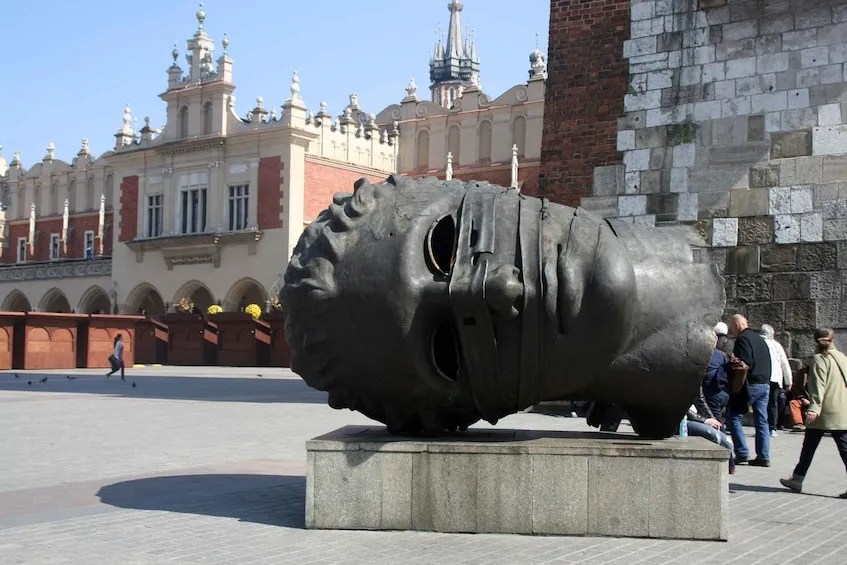 A large metal sculpture of a distorted human head placed on a stone pedestal, located in a public square. The background features historic buildings and a crowd of people walking by.