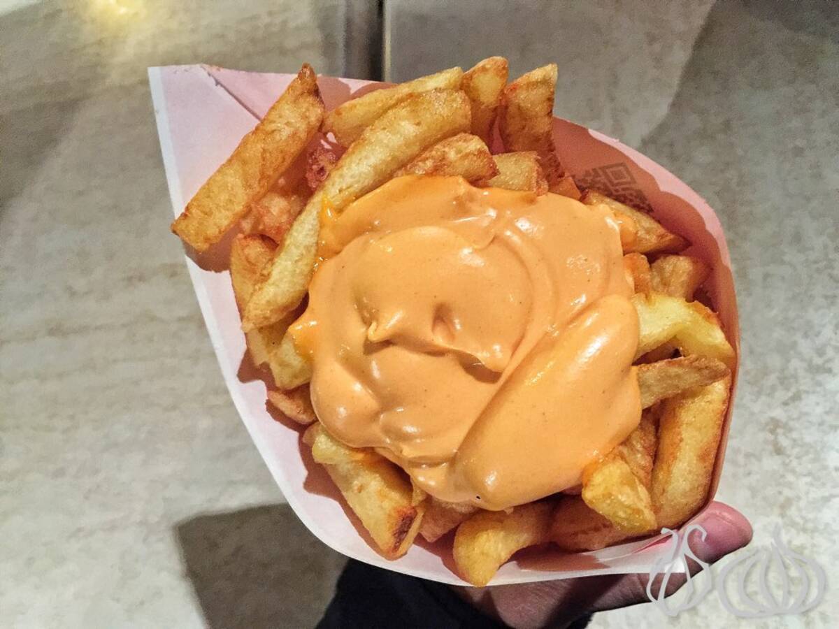 A close-up of crispy French fries topped with a creamy orange sauce, served in a paper cone.