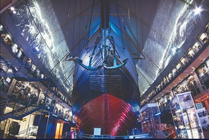 A striking interior view of the Viking Ship Museum, featuring a large Viking ship illuminated in blue and red light, with exhibition displays and walls showcasing maritime history.