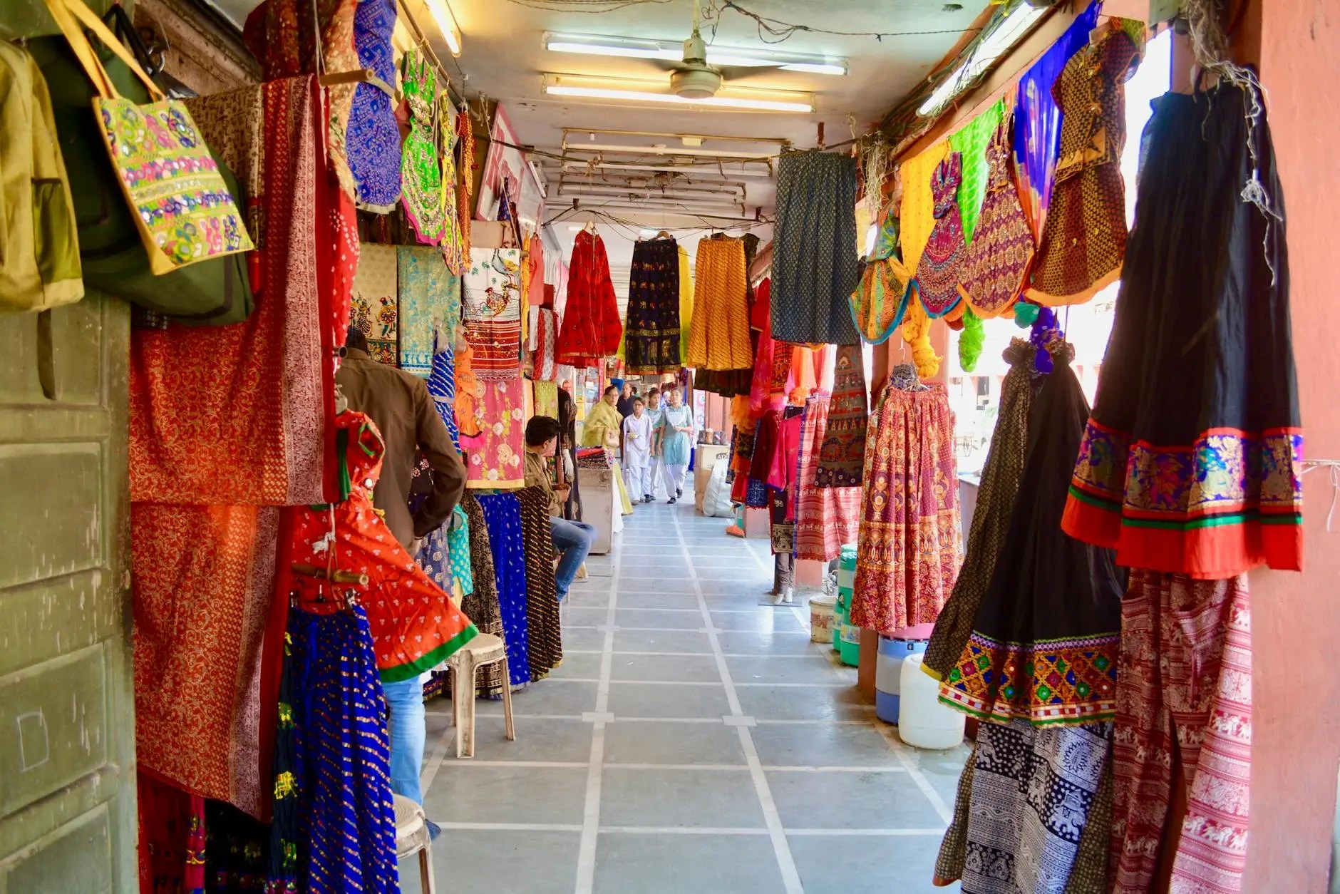 A vibrant market alley in Jaipur, showcasing colorful traditional clothing and textiles hanging from the walls, with shoppers visible in the background.