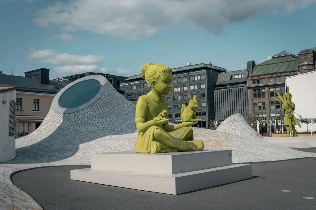 A bright green statue of a seated child holding a rabbit, located in Lasipalatsi Square, Helsinki, with modern architectural elements in the background.