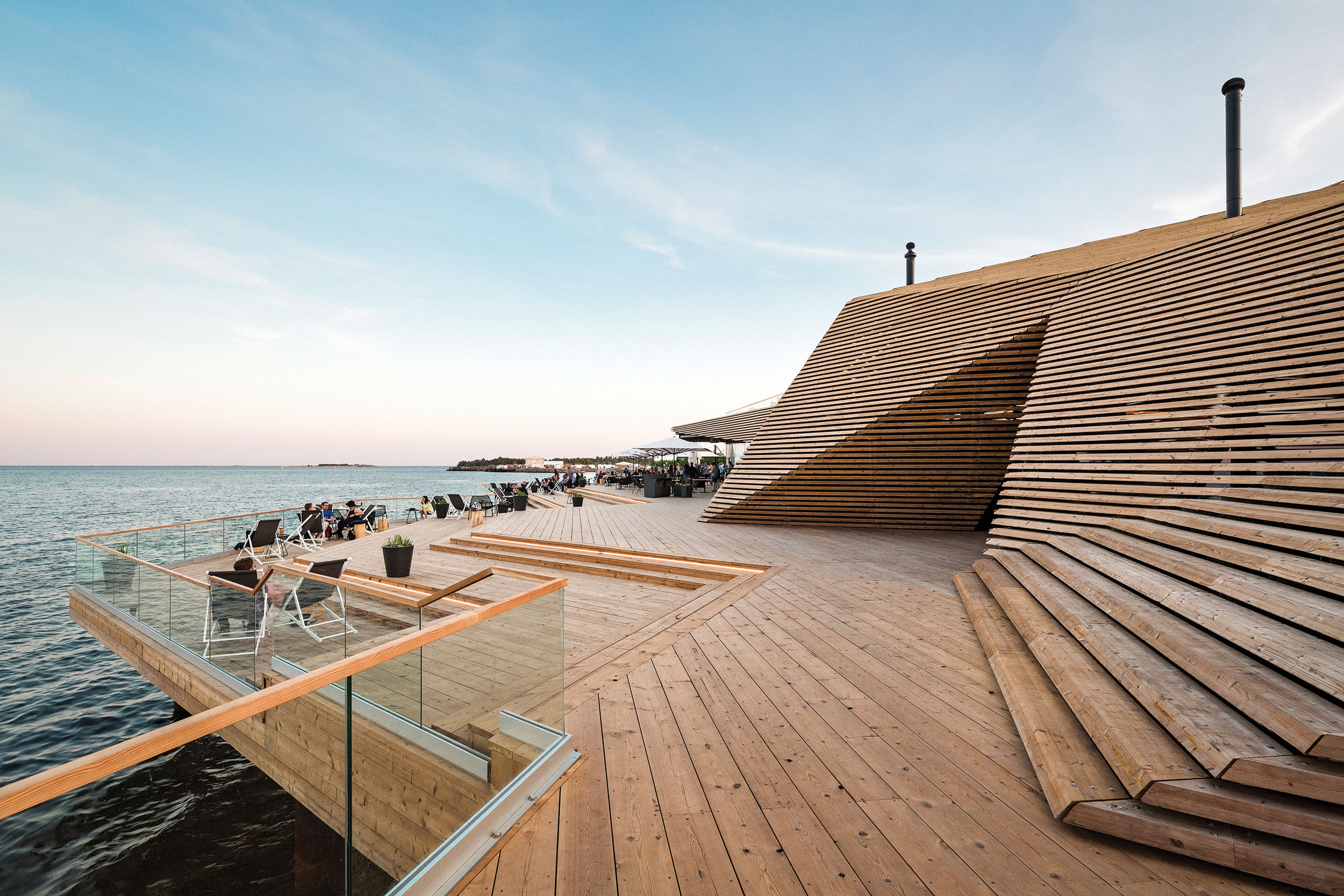 A wooden terrace overlooking the sea, designed with sloped sections and glass railings. Visitors relax on lounge chairs as they enjoy the coastal view.