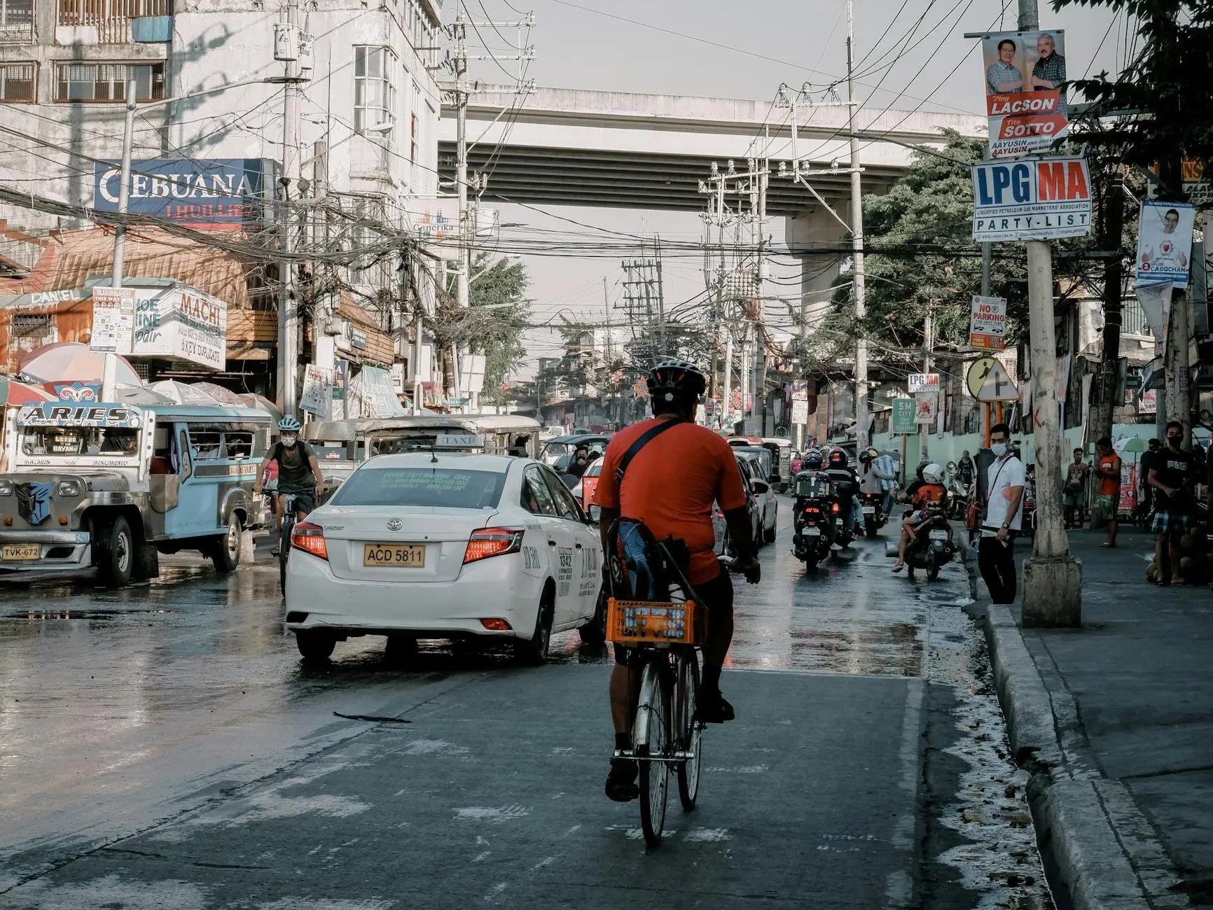 A cyclist riding through a busy street in Manila, surrounded by traffic including cars and motorbikes, with urban buildings and power lines in the background.