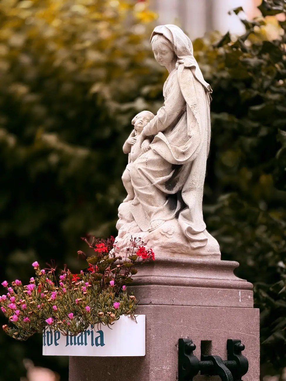 A stone statue of a woman with a child, surrounded by colorful flowers in a planter, with a sign that reads 'de Maria'.