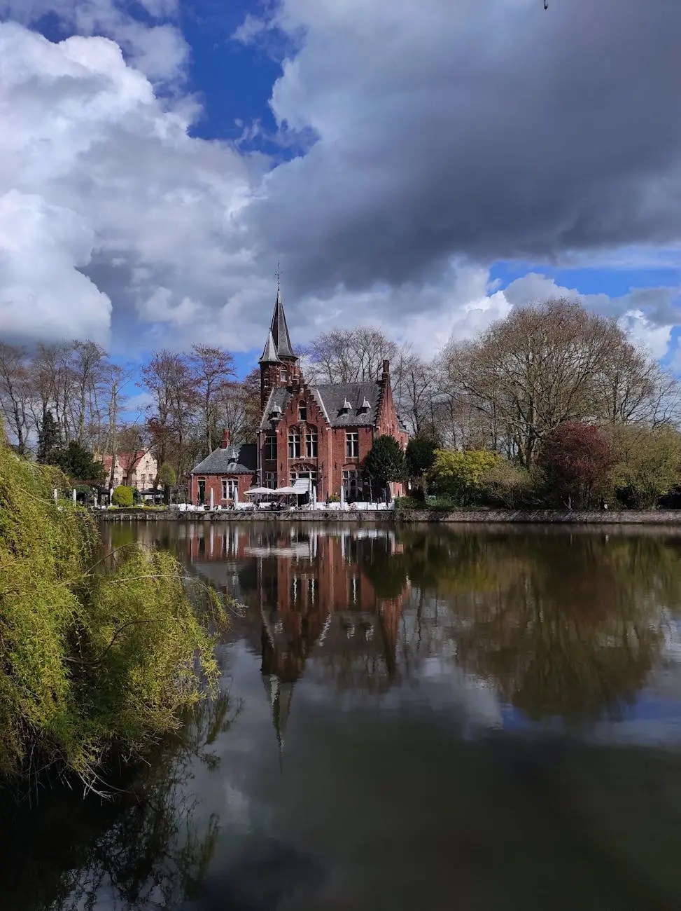 A picturesque view of a red-bricked building with a tall spire, surrounded by lush trees and reflecting in a calm lake under a partly cloudy sky in Bruges, Belgium.