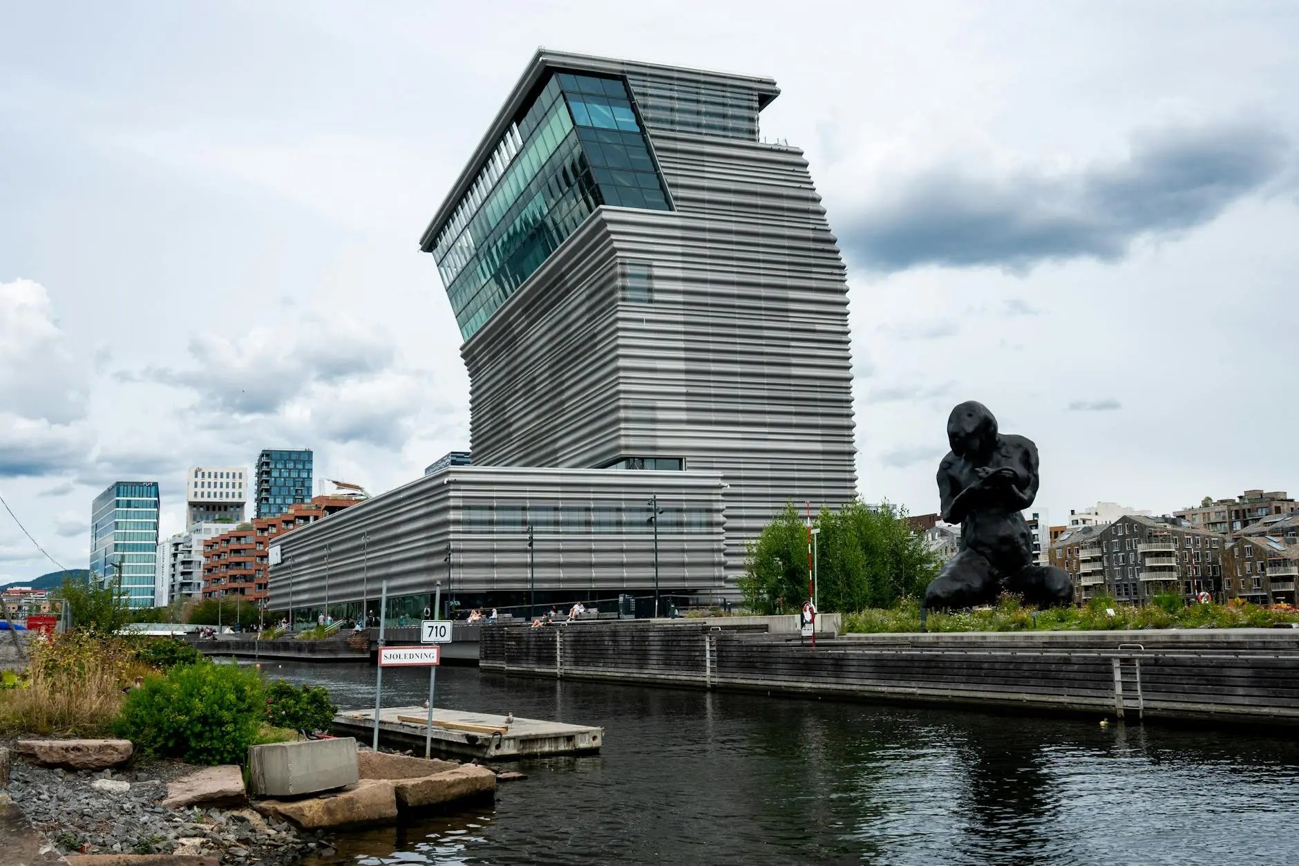 A modern architectural building with a unique design stands next to a waterfront in Oslo, Norway, featuring jagged lines and glass elements, accompanied by a large black sculpture nearby.