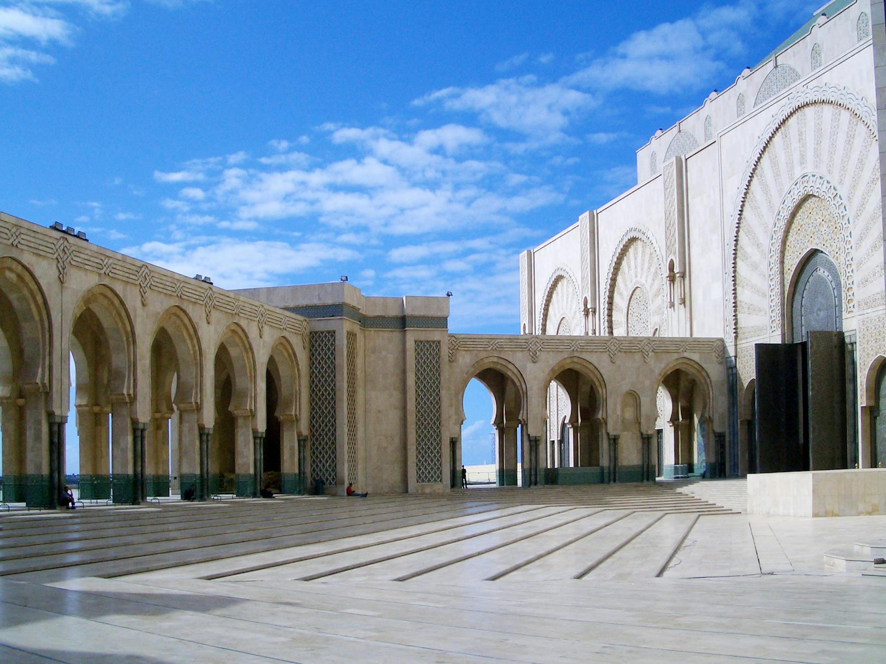 The entrance courtyard of the Hassan II Mosque in Casablanca, featuring ornate arches and intricate tile work under a blue sky.