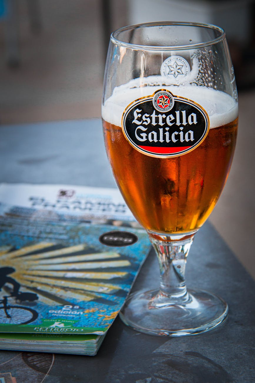 A glass of Estrella Galicia beer resting on a table with a colorful brochure in the background.
