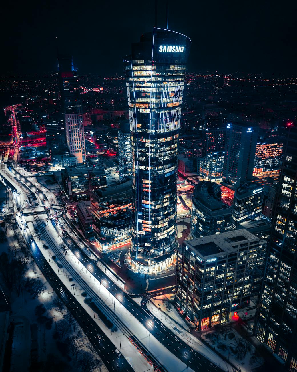 Aerial view of a modern cityscape in Warsaw at night, featuring the illuminated Samsung building and a busy street with vehicles and bright lights.