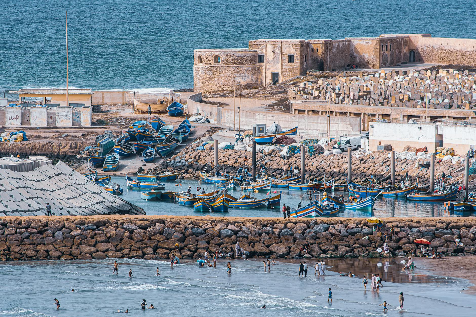 A scenic view of a coastal area in Casablanca, featuring colorful fishing boats docked near a rocky shoreline, people enjoying the beach, and an old building in the background against a serene ocean backdrop.
