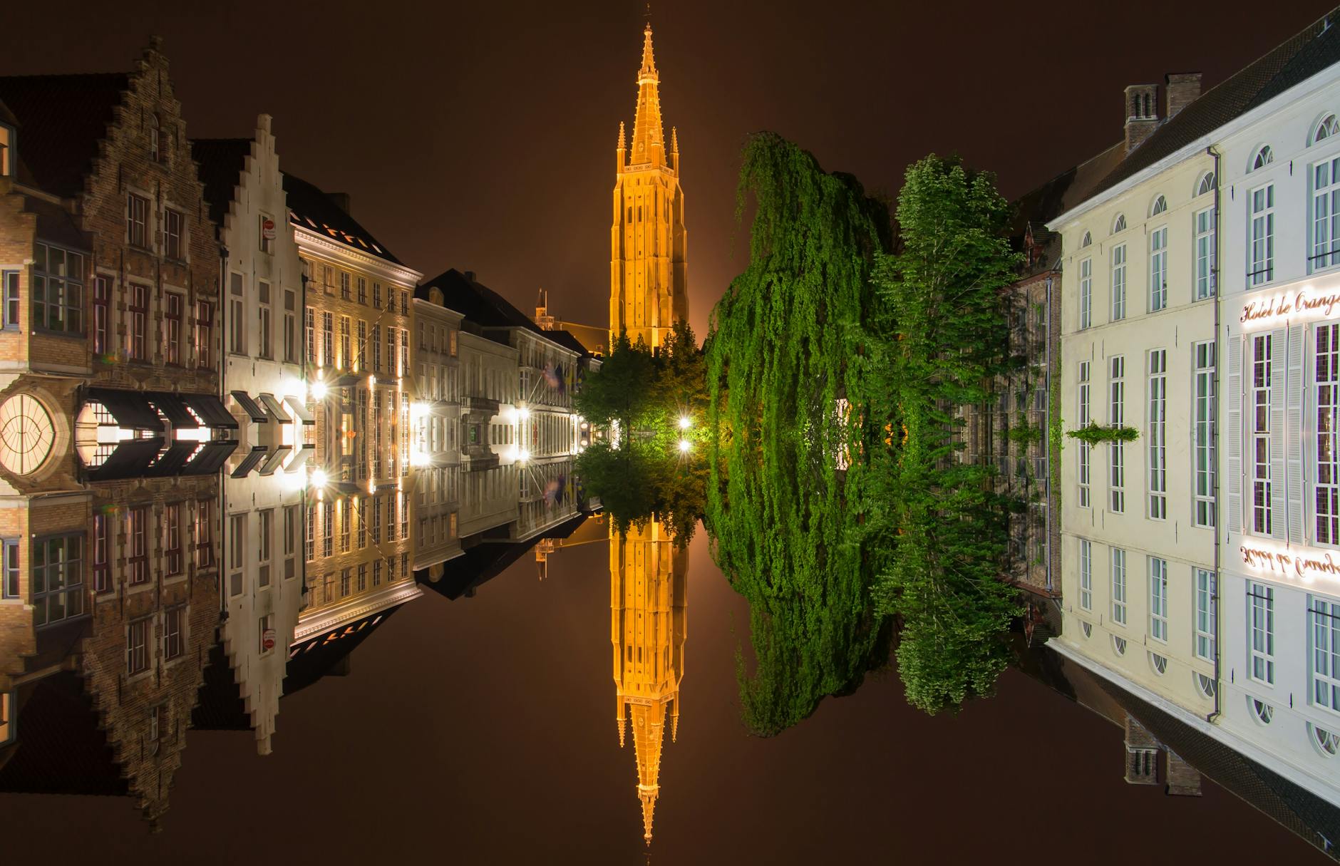 Night view of Bruges reflecting in a canal, featuring illuminated buildings and the Belfry tower.
