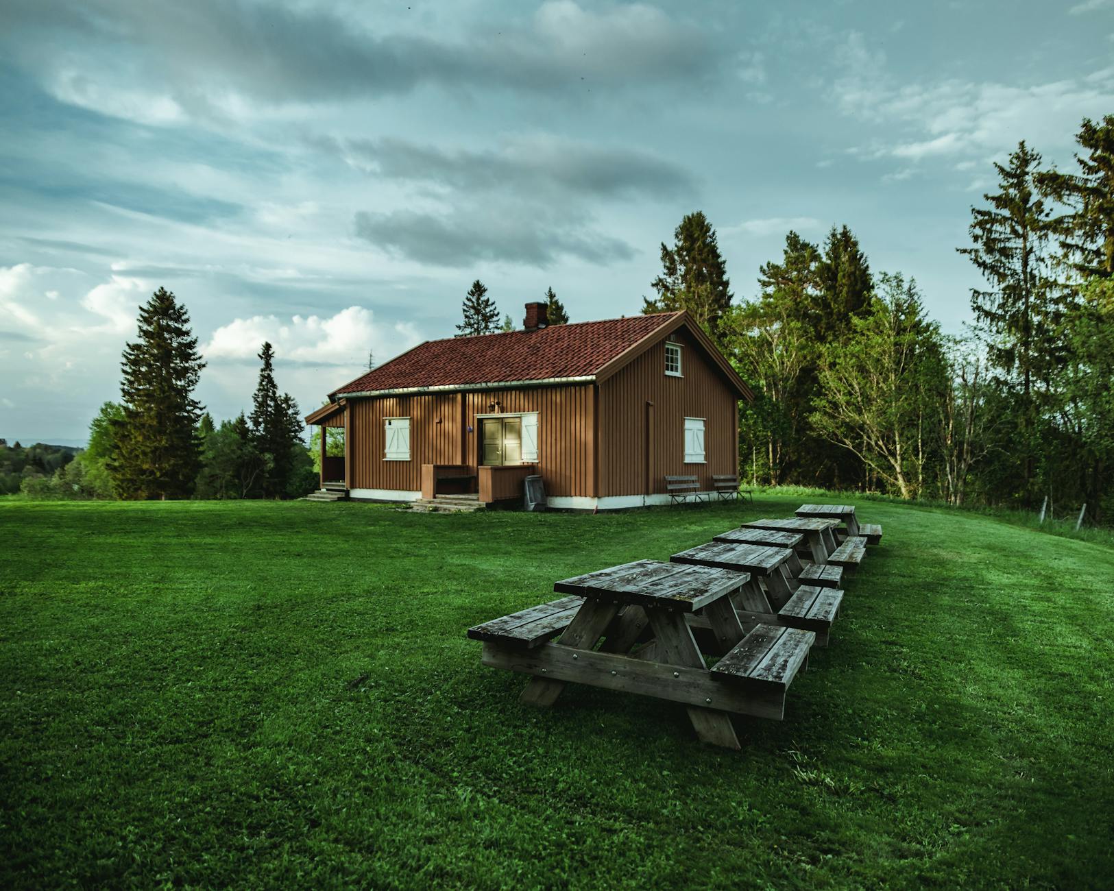 A cozy wooden house with a red roof set in a lush green landscape, featuring picnic tables arranged on the grass, surrounded by tall trees under a partly cloudy sky.