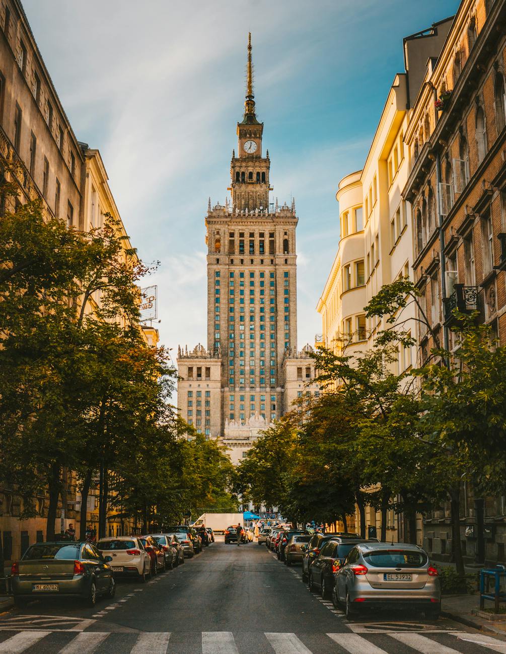 A street view in Warsaw showcasing the iconic Palace of Culture and Science, a tall, ornate building, surrounded by trees and parked cars under a clear blue sky.