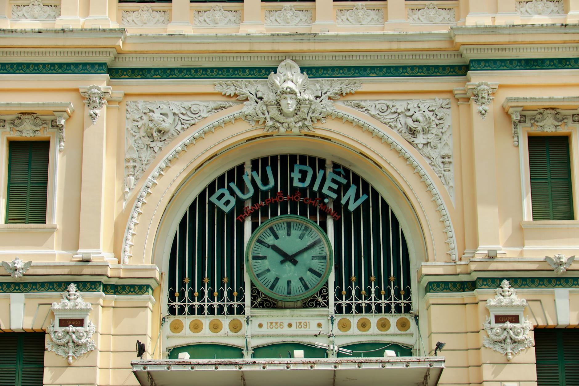 Close-up view of the ornate facade of the Saigon Central Post Office, featuring a large clock and decorative architectural details, with the words 'BƯU ĐIỆN' prominently displayed.