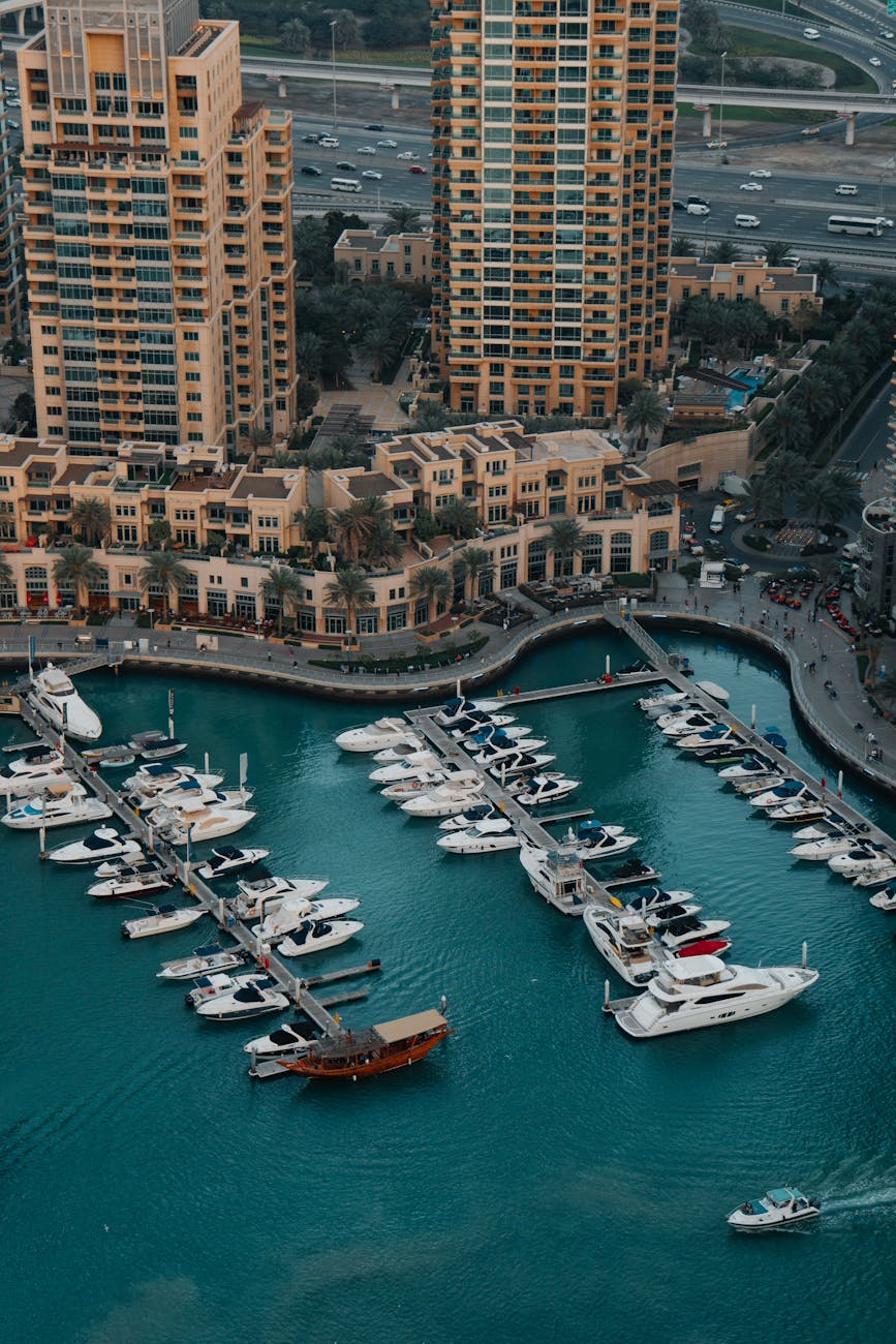 An aerial view of a marina in Doha, Qatar, showcasing numerous luxury yachts docked in vibrant blue waters, surrounded by modern high-rise buildings and palm trees.
