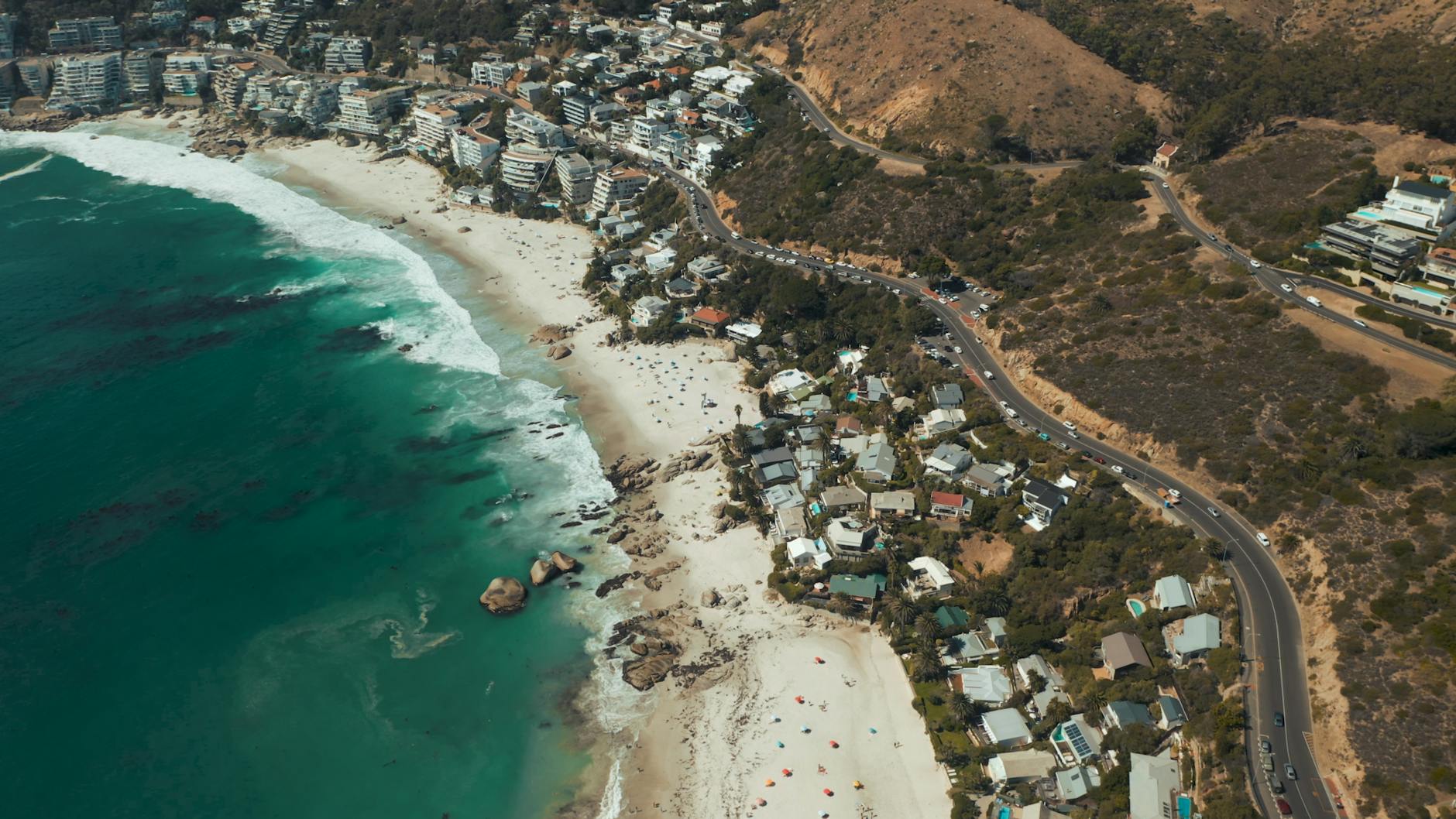 Aerial view of a picturesque beach with clear turquoise waters, sandy shorelines, and scattered beachgoers, surrounded by residential buildings and mountainous terrain.