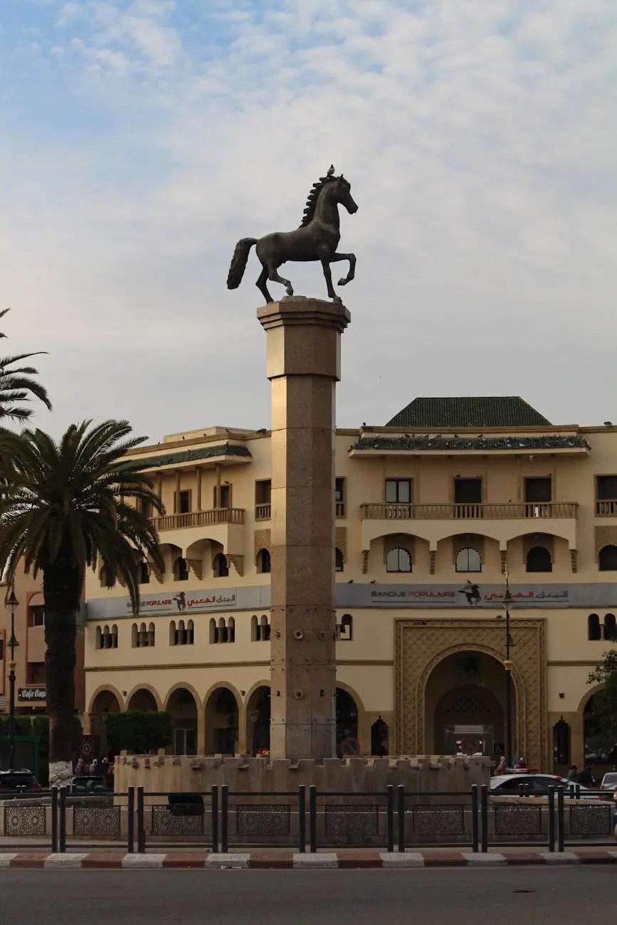 Statue of a horse atop a tall pedestal in a public square, with palm trees and buildings in the background under a cloudy sky.