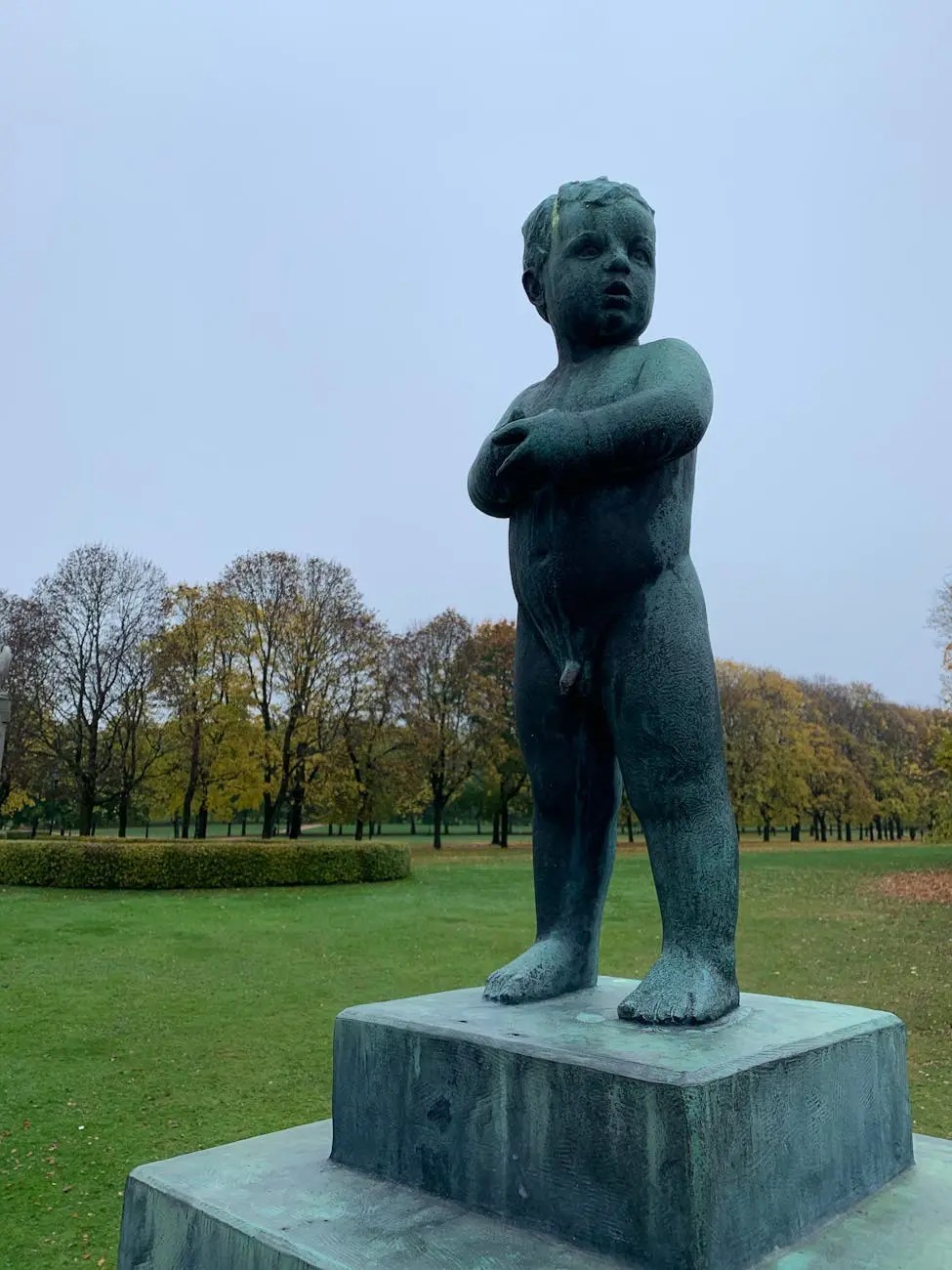 A bronze statue of a small child with crossed arms, standing on a stone pedestal, surrounded by green grass and trees in the background.