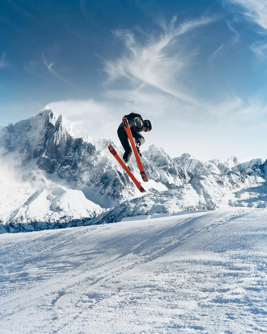 A skier performing a jump against a backdrop of majestic snow-capped mountains and a clear blue sky.