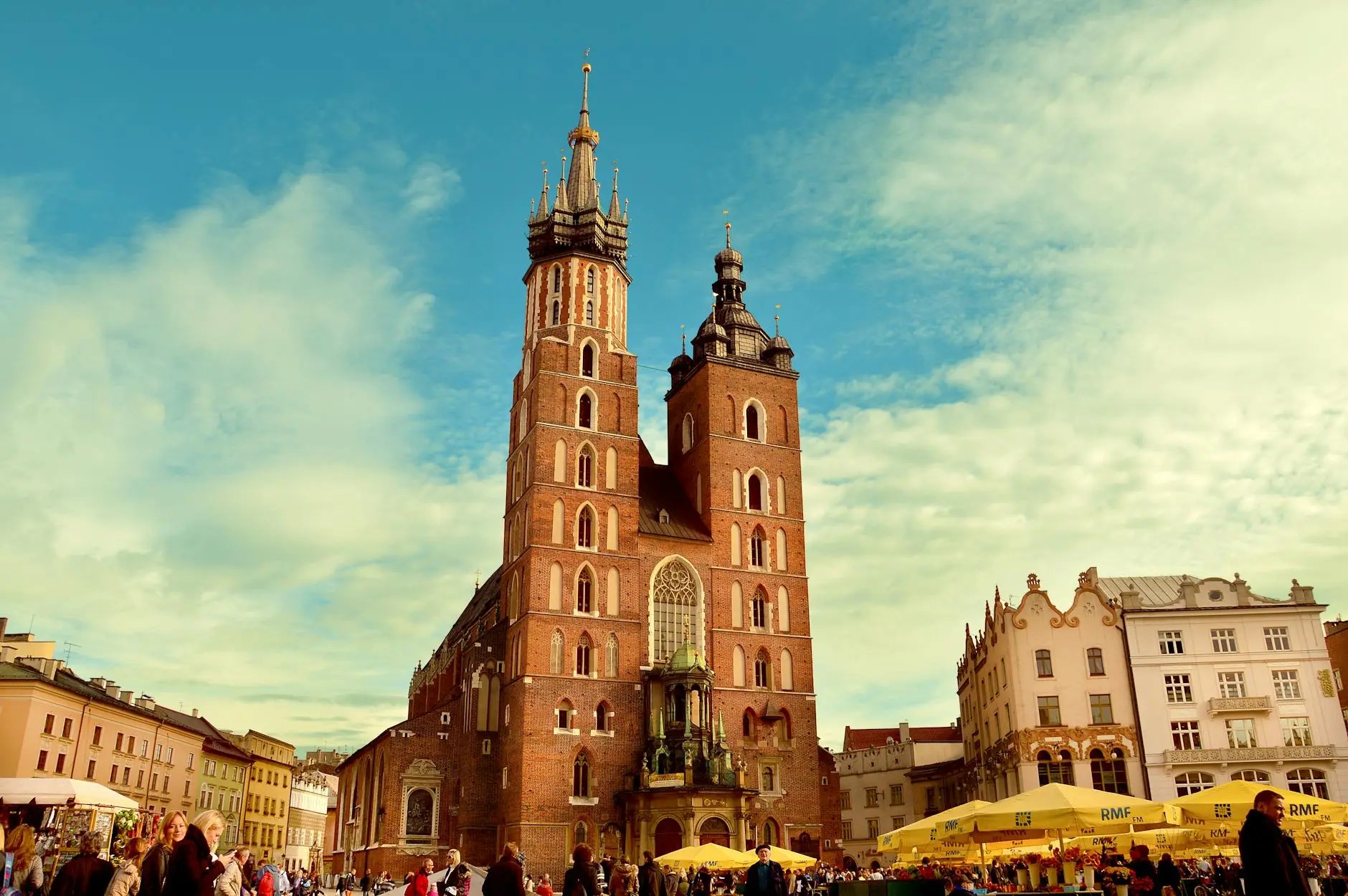 A view of St. Mary's Basilica in Kraków, Poland, with its two distinct towers and historic architecture, surrounded by a bustling market square filled with people.