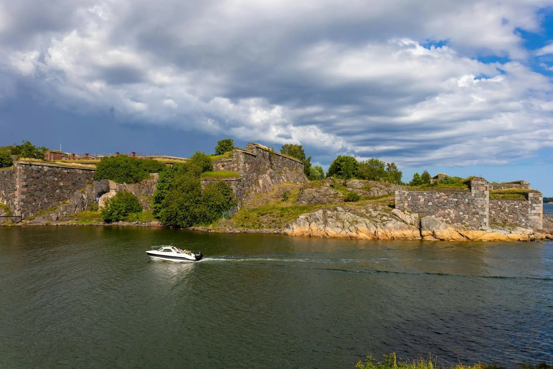 A view of the Suomenlinna Sea Fortress in Helsinki, Finland, with a boat passing by on the water and a cloudy sky above.