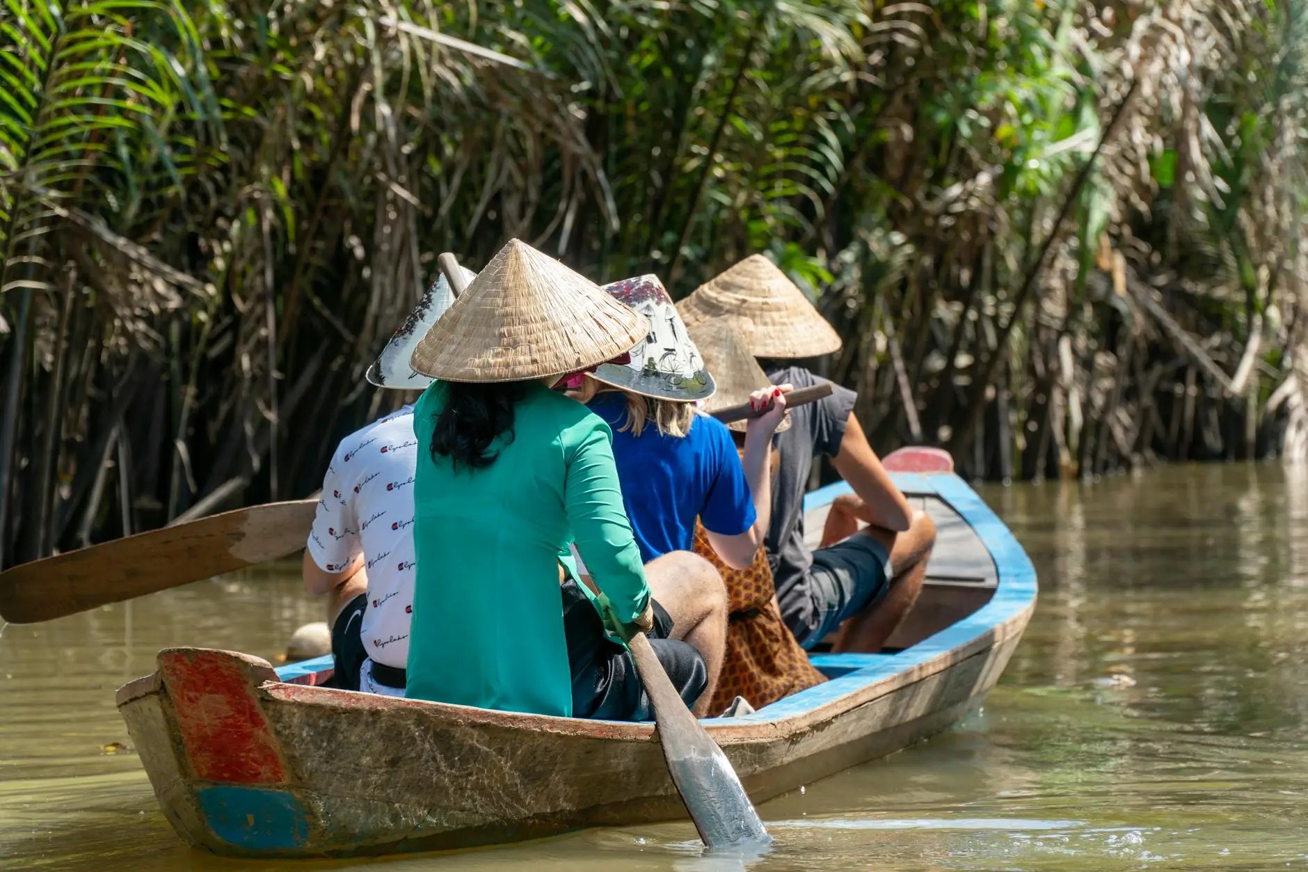 A group of people wearing traditional conical hats, paddling a small boat through a river surrounded by lush greenery.