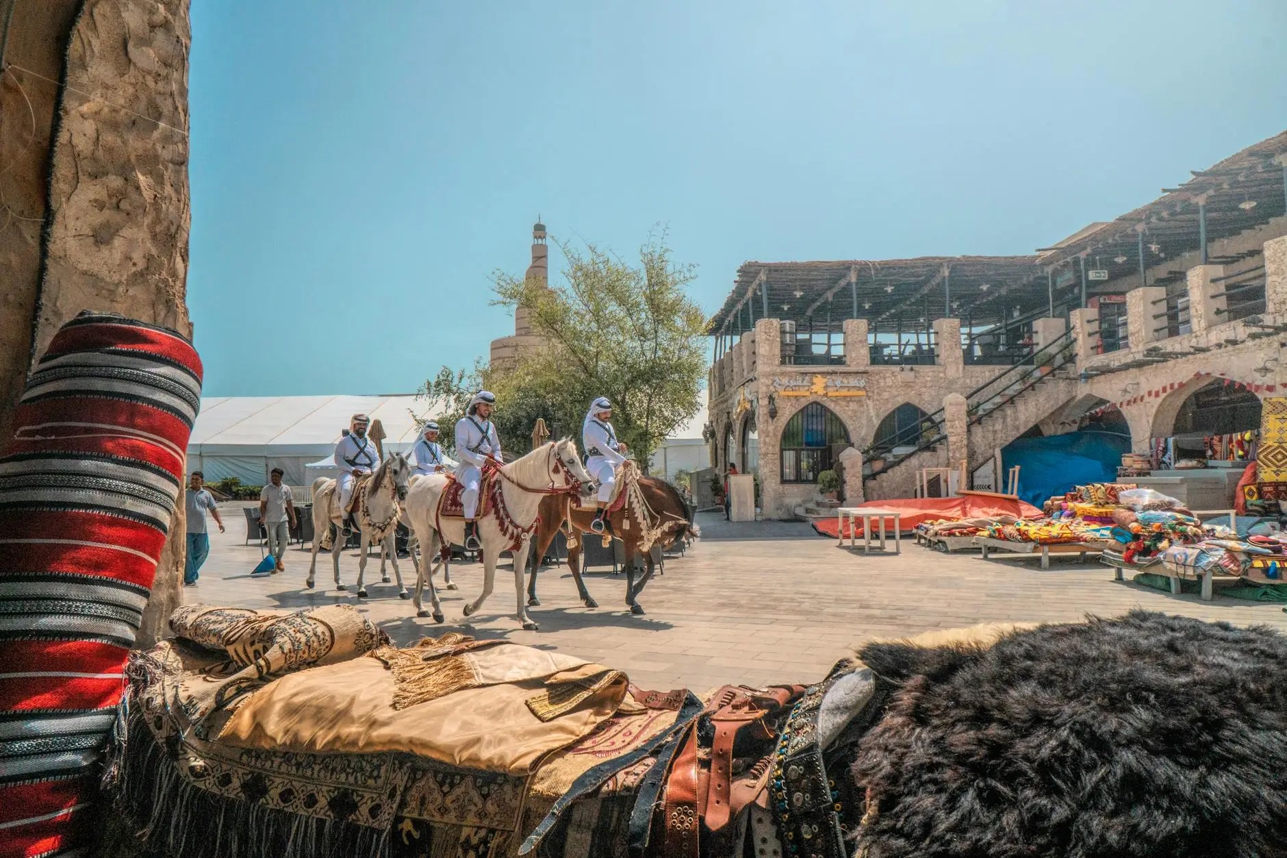 A bustling scene at Souq Waqif in Doha, Qatar, featuring men on horseback in traditional attire, surrounded by colorful market stalls and woven textiles.