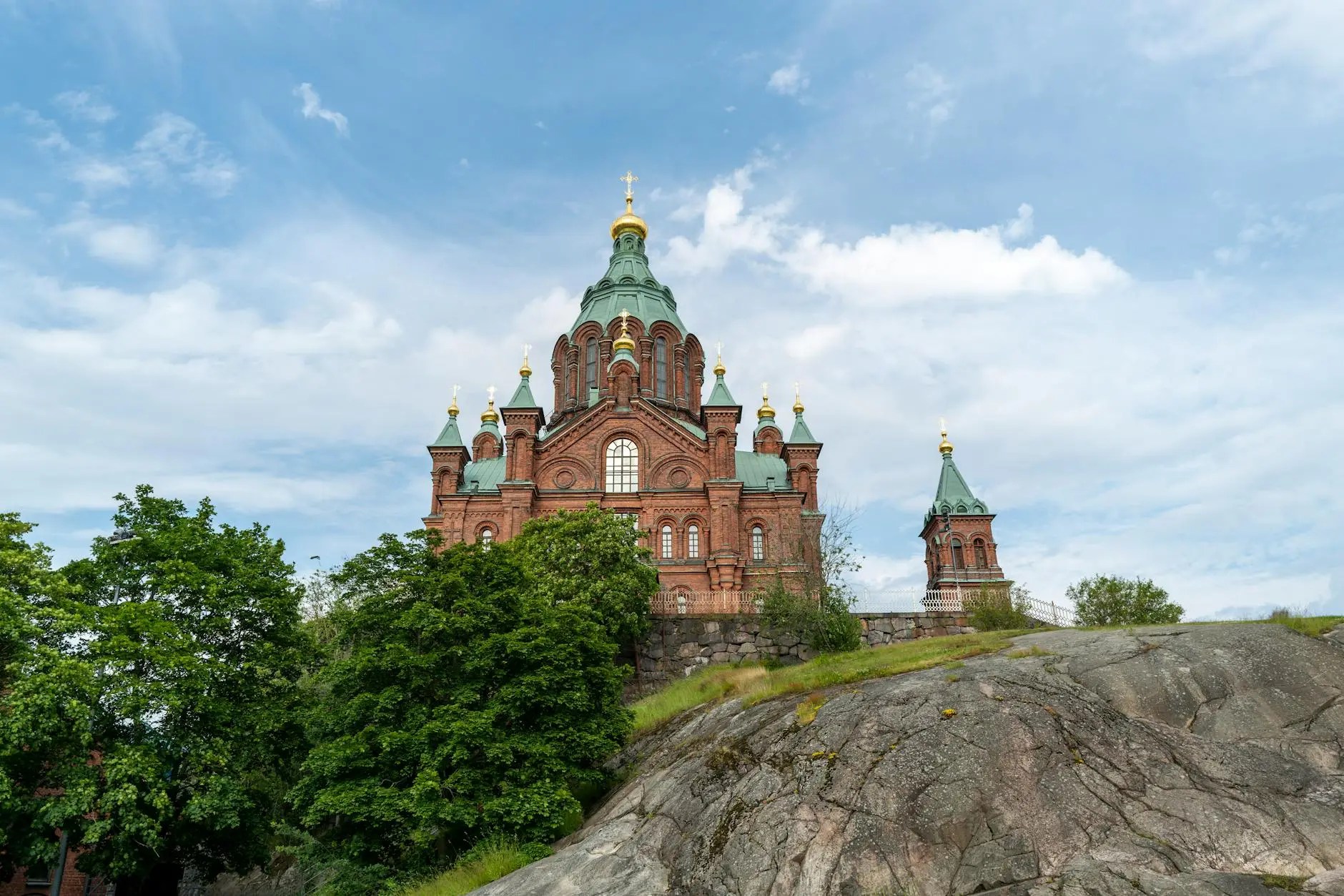 View of the Uspenski Cathedral in Helsinki, featuring its distinctive red brick architecture and green domes against a blue sky with scattered clouds.