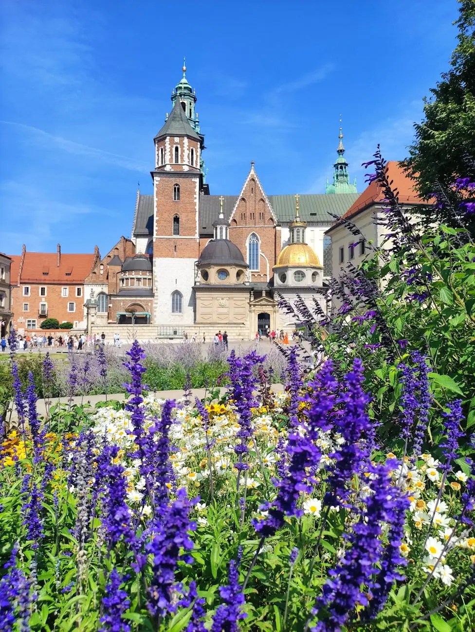 A vibrant view of colorful flowers in the foreground with the historic architecture of Wawel Castle in Kraków, Poland, under a clear blue sky.