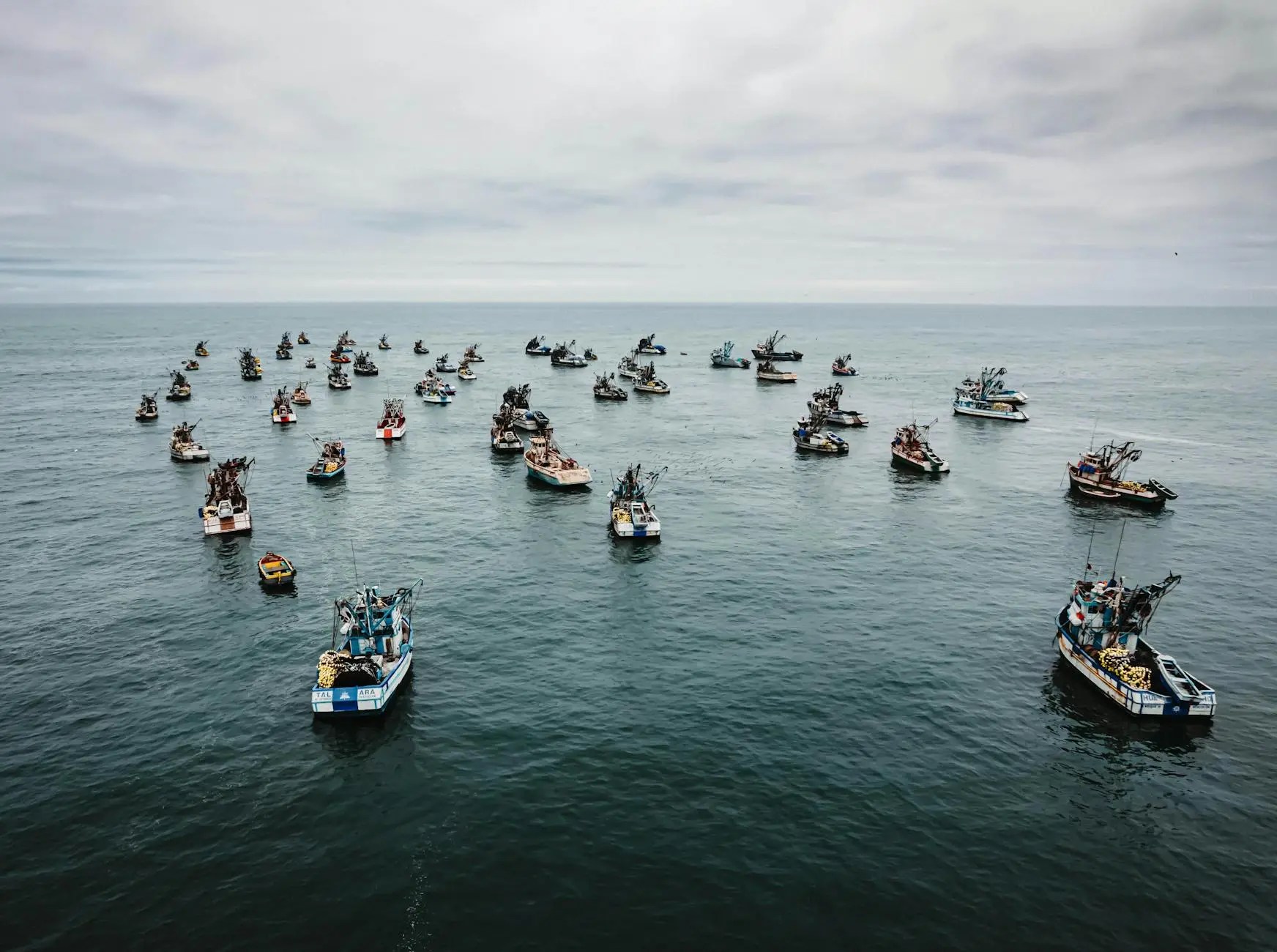 Aerial view of numerous fishing boats scattered in the ocean off the coast of Lima, Peru, under a cloudy sky.