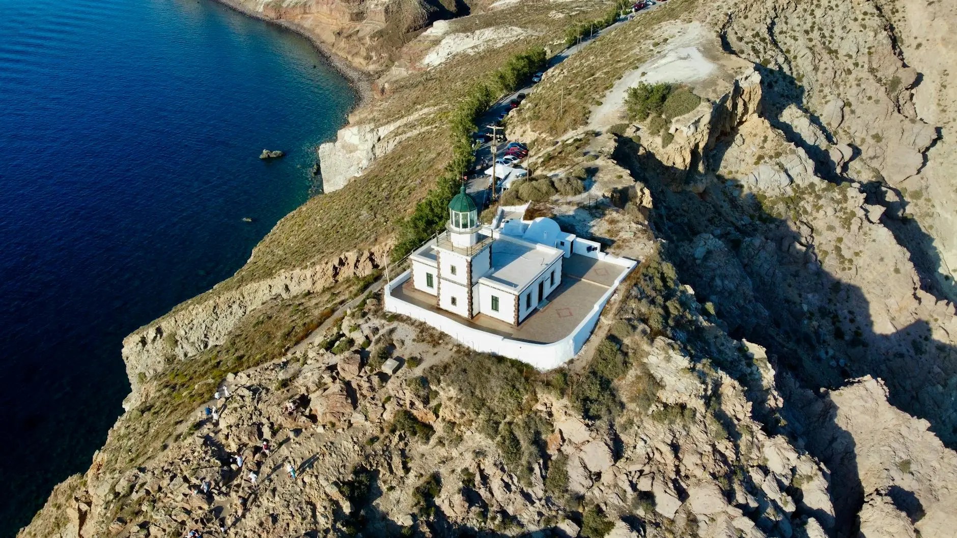 Aerial view of a lighthouse situated on a rocky cliff overlooking a calm blue sea, surrounded by rugged terrain.