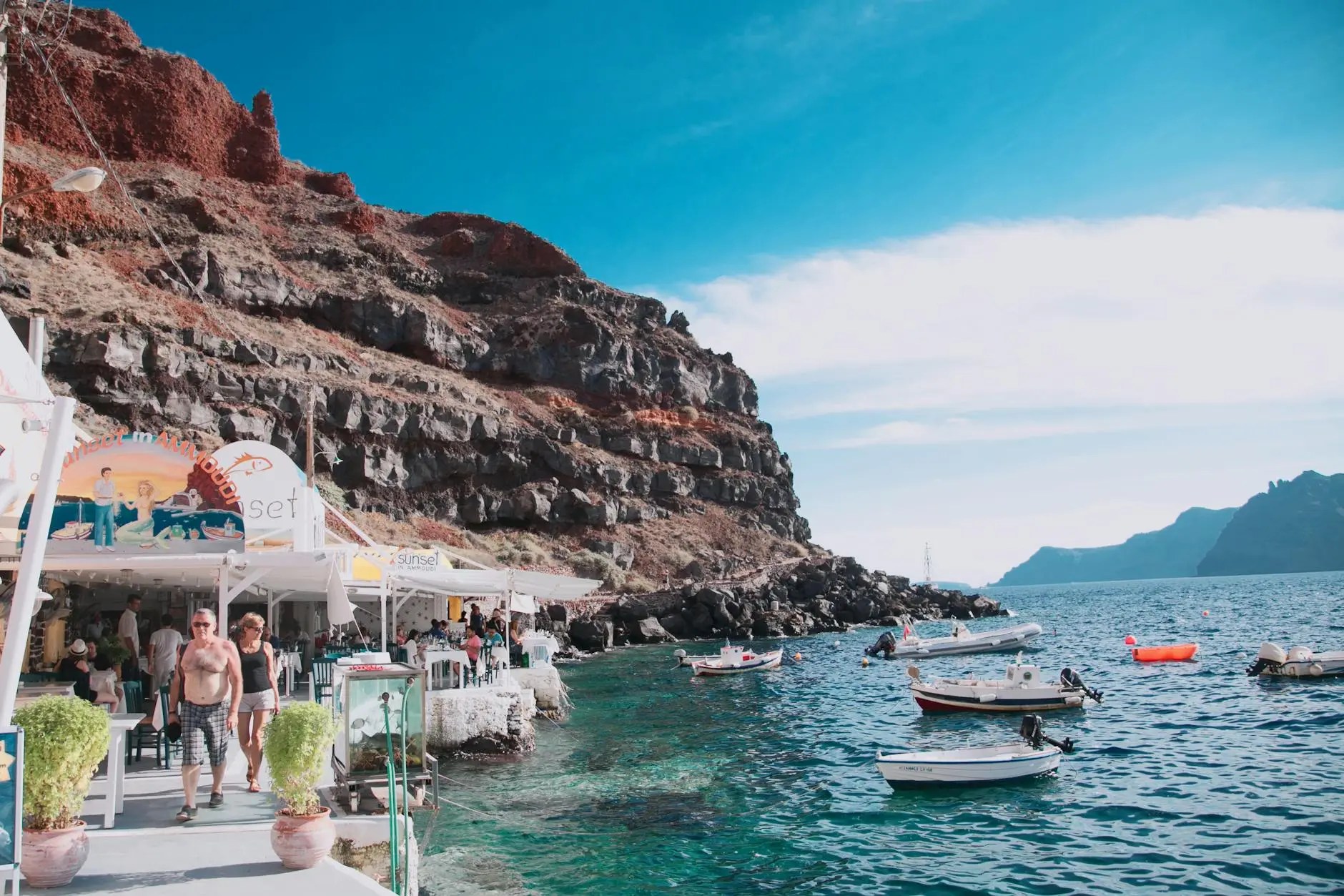 View of a picturesque coastal area in Santorini with restaurants along the shoreline, surrounded by rocky cliffs and boats floating in the clear water under a bright blue sky.