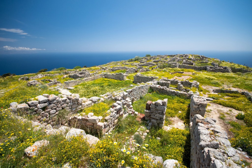 Ruins of an ancient settlement on a hillside covered in green grass and wildflowers, overlooking the sea under a clear blue sky.