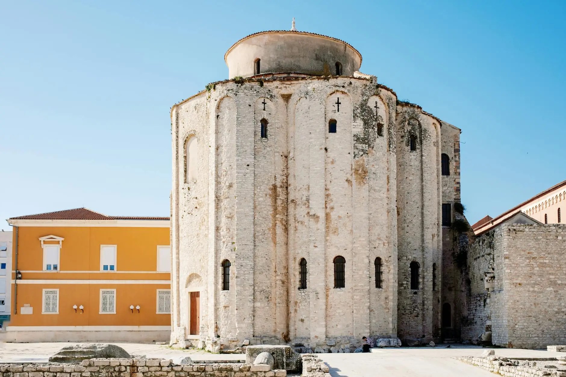A historic round stone building with a domed roof and arched windows, surrounded by a mix of modern and ancient architecture in Zadar, Croatia.