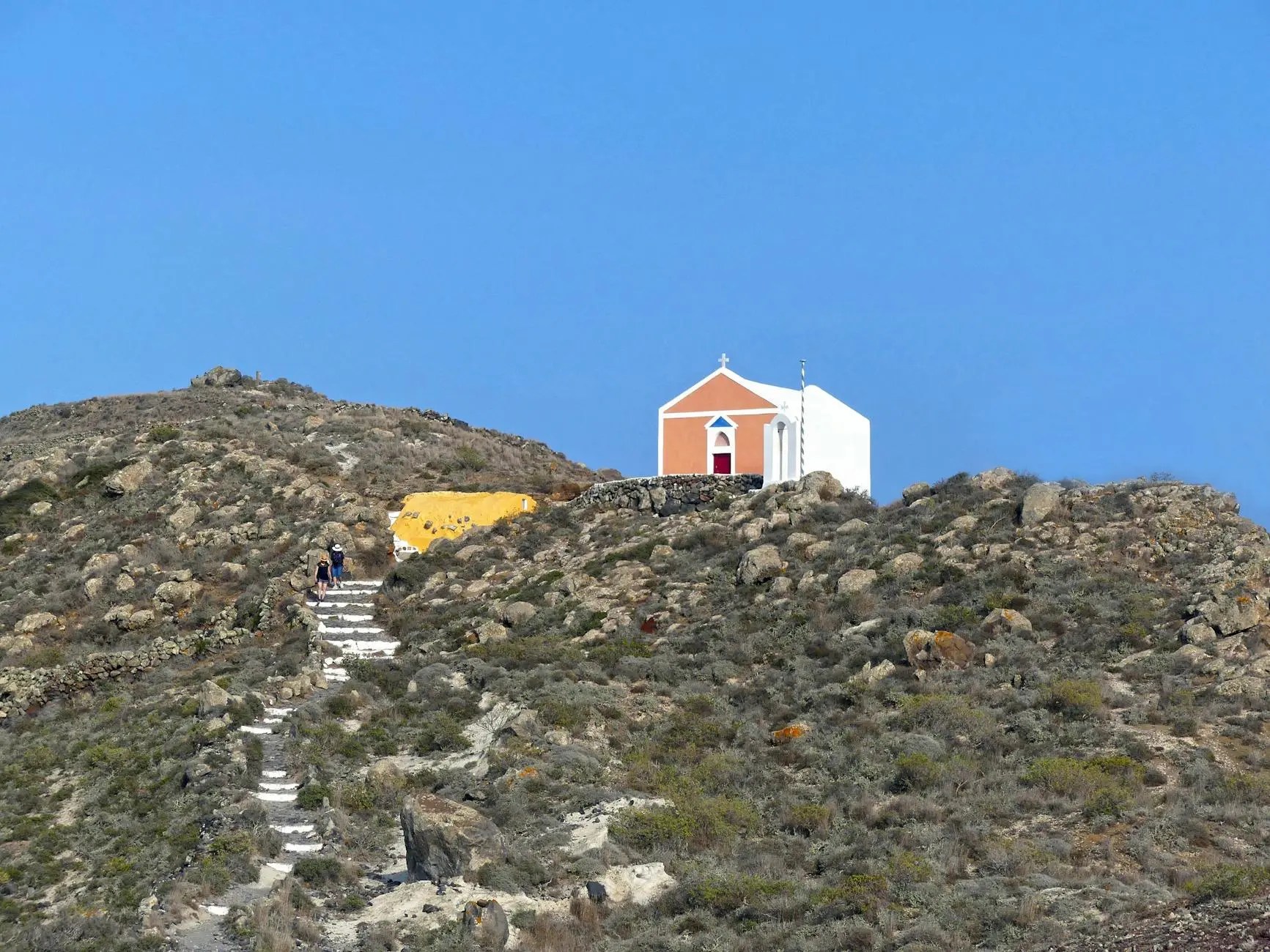 A picturesque hillside approach to a small church with a pink and white facade, situated on a rocky terrain beneath a clear blue sky.