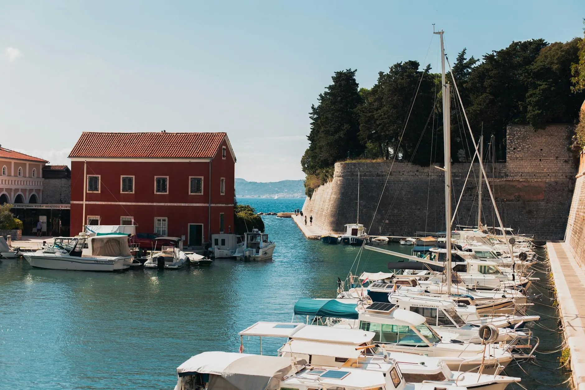 A scenic view of a marina in Zadar, Croatia, featuring a variety of boats moored in the water, with a red building and green trees in the background under a clear blue sky.