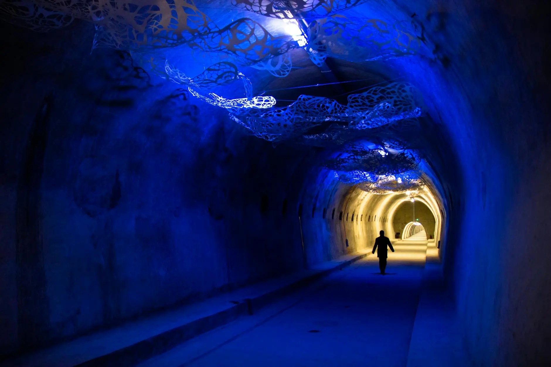 A person walking through a dimly lit tunnel adorned with blue lights and intricate paper-like decorations on the ceiling.