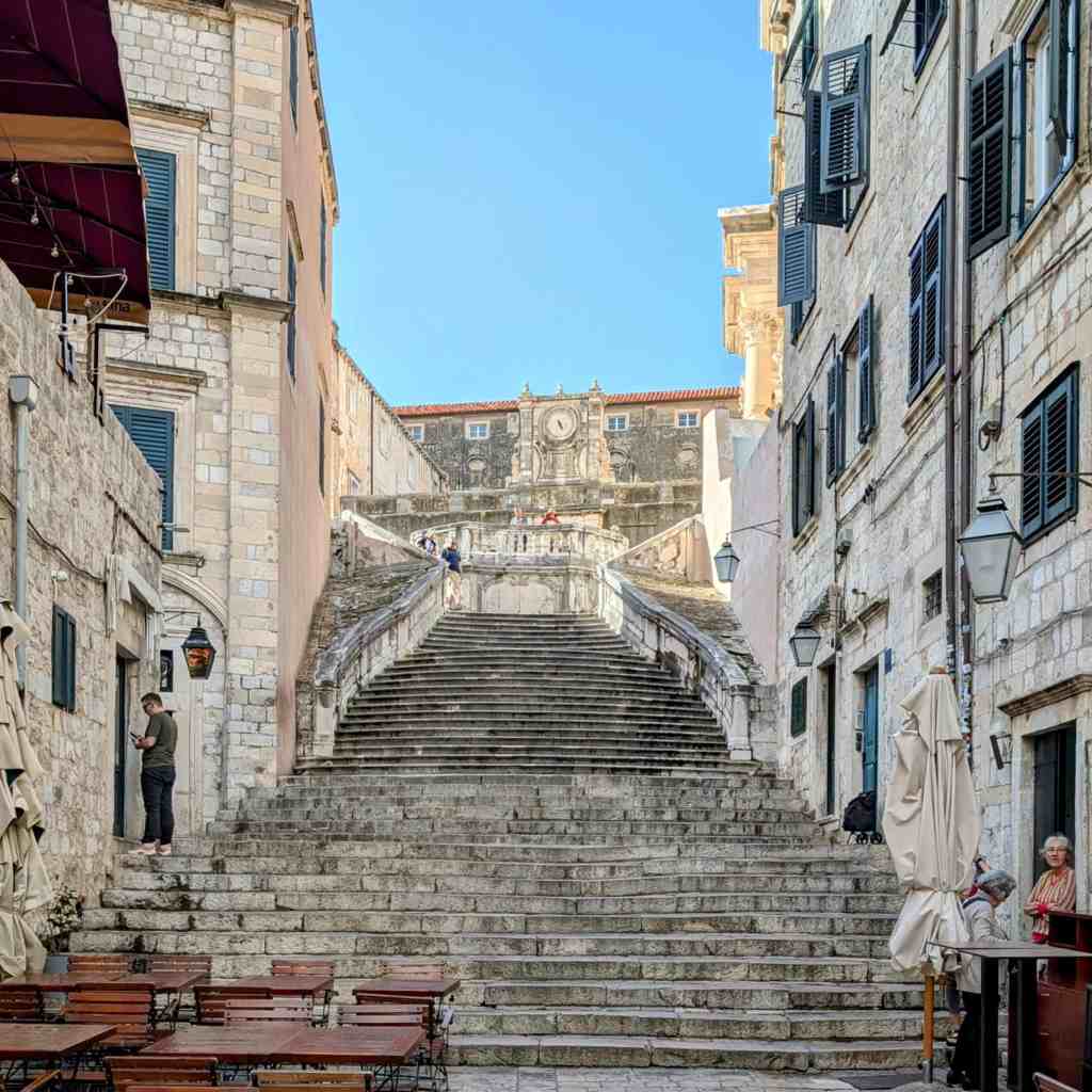 A view of a historic staircase in Dubrovnik, flanked by stone buildings and decorative lanterns, leading toward a clock tower in the background.