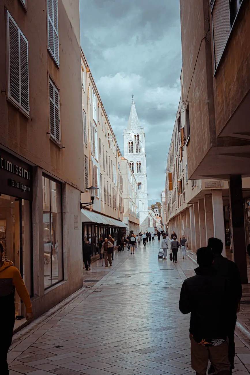 A bustling street in Zadar, Croatia, lined with shops and cafes, featuring a glimpse of a historic bell tower in the background under a cloudy sky.