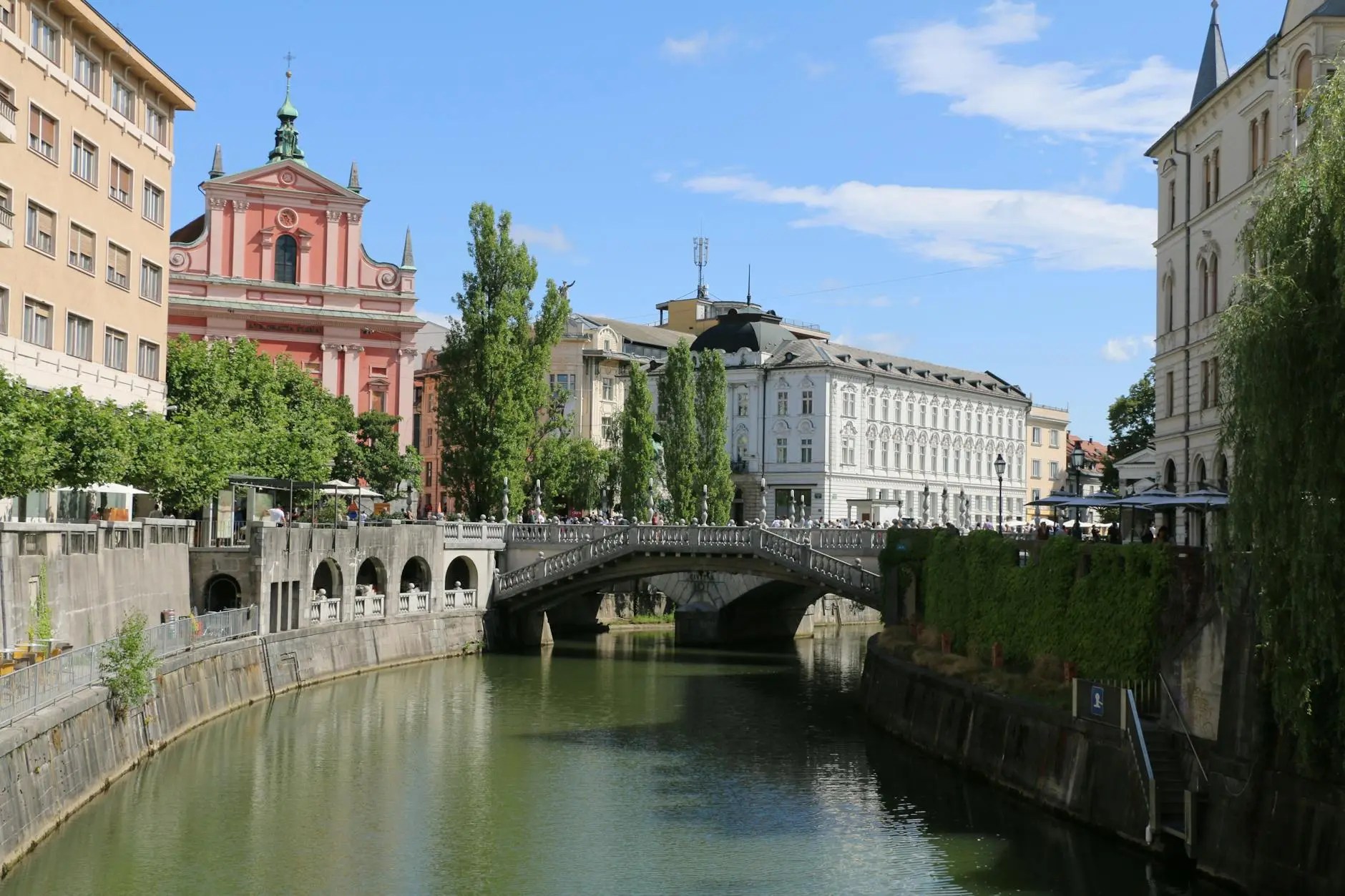 View of the Ljubljanica River with a bridge and cafes along the banks, framed by colorful pastel buildings and greenery in Ljubljana, Slovenia.