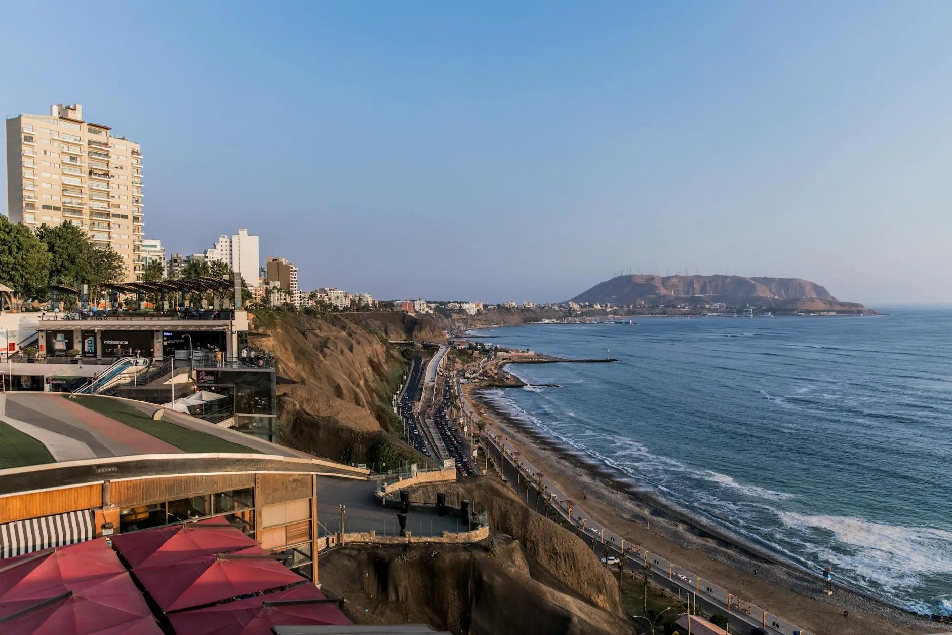 A panoramic view of the coastline in Lima, Peru, featuring a cliffside park, modern buildings, and the Pacific Ocean under a clear blue sky.