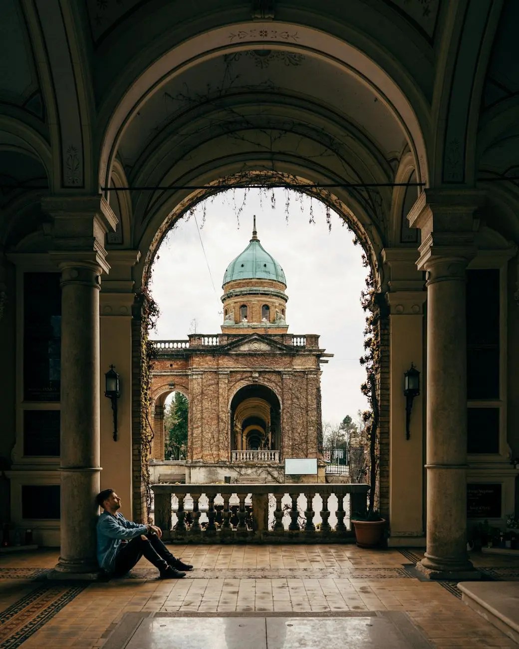 A person sitting against a wall in a historic building, looking contemplative towards a view of an ornate dome and arched colonnades.