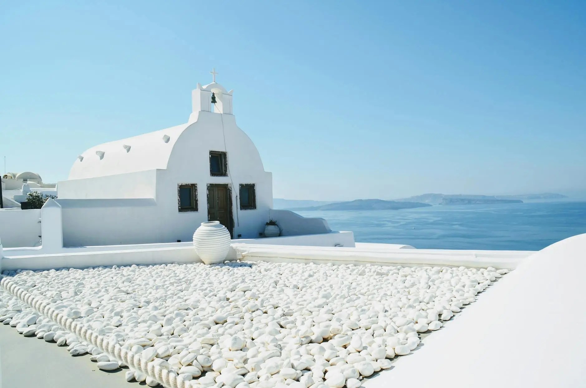 A view of a white dome building in Santorini, Greece, with a blue sky and the Aegean Sea in the background, featuring white pebbles in the foreground.