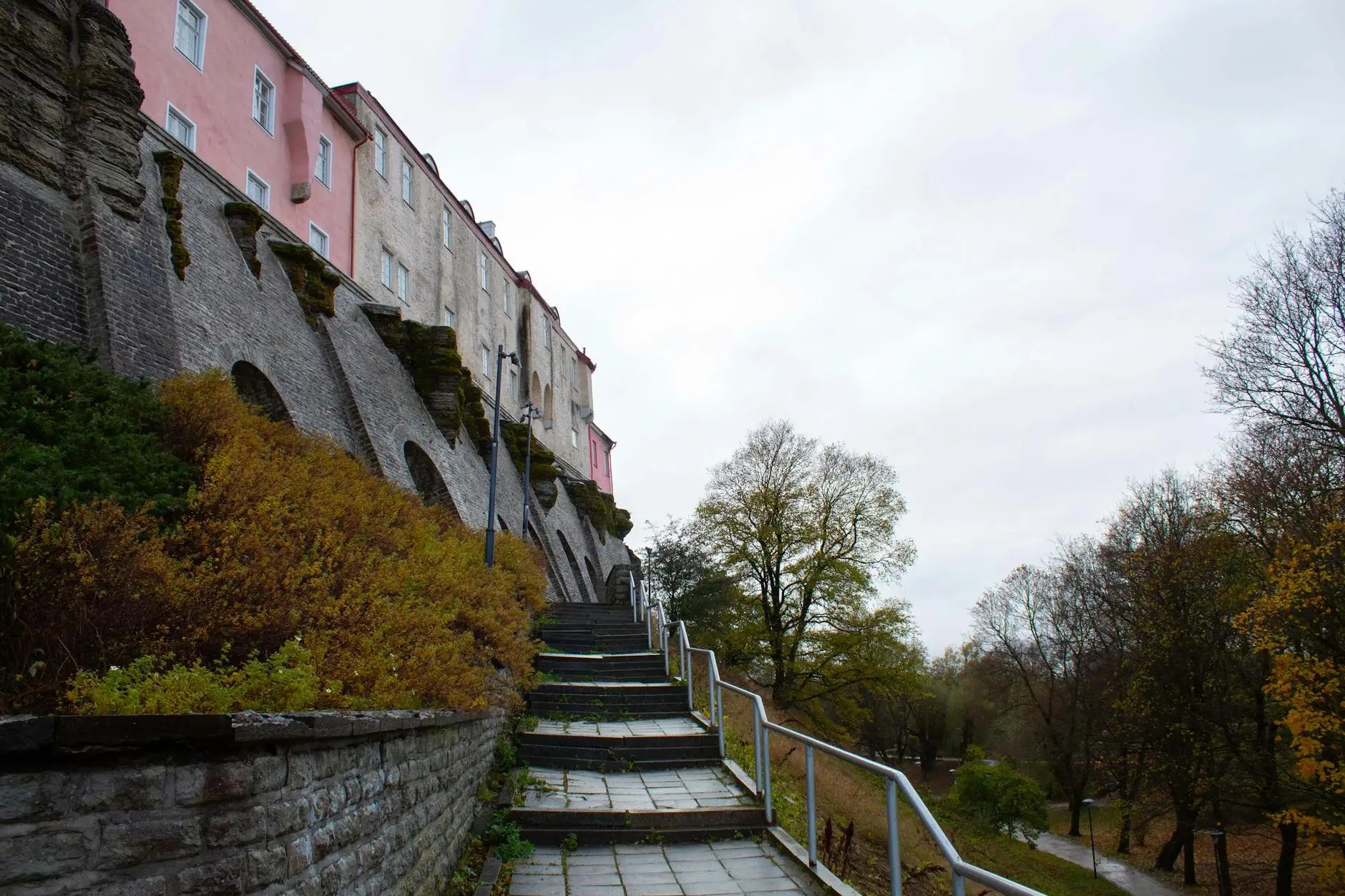 A stairway leading up to pastel-colored buildings and a stone wall overgrown with greenery, set against a cloudy sky.
