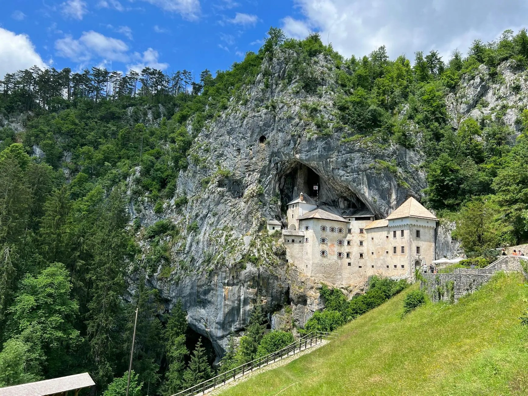 A stunning view of Predjama Castle, built into a cliffside, surrounded by dense green forest and dramatic rock formations under a bright blue sky.